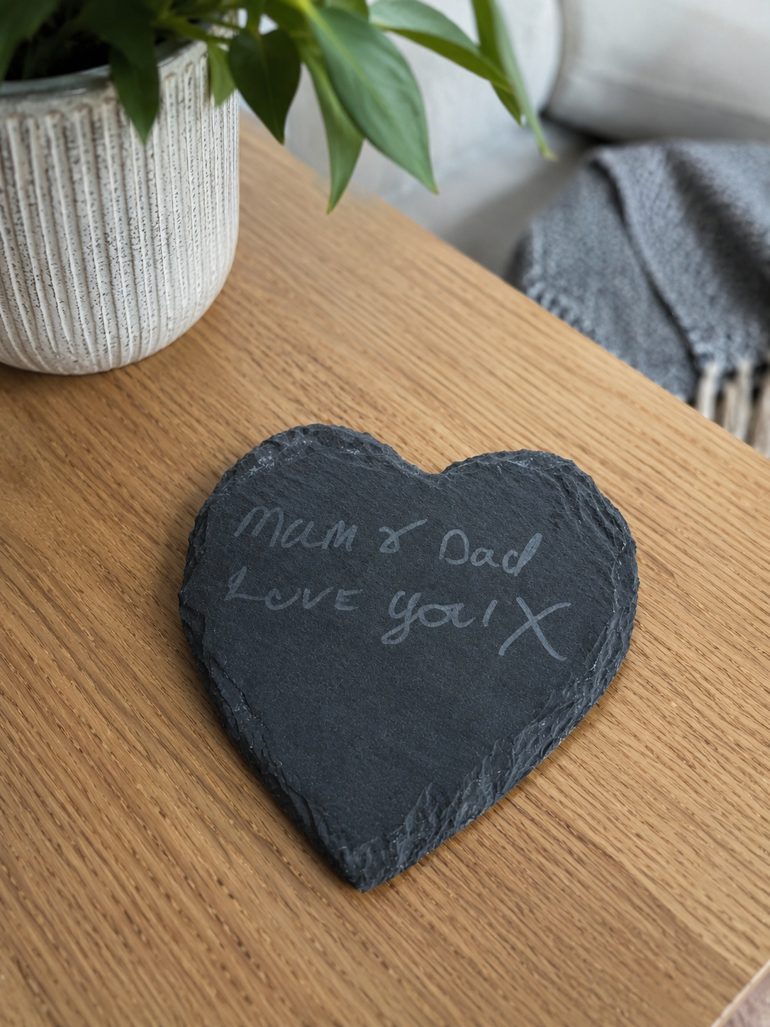 Heart-shaped slate with 'Mum & Dad Love you!' on a wooden surface with a plant and scarf in the background.