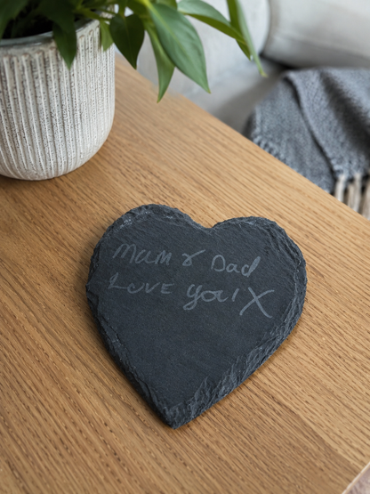 Heart-shaped slate with 'Mum & Dad Love you!' on a wooden surface with a plant and scarf in the background.