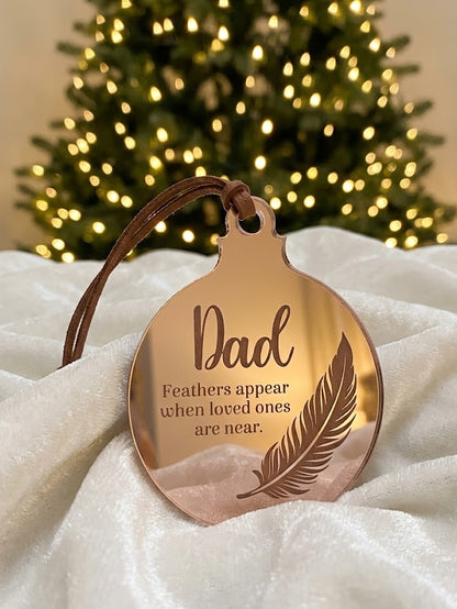 A round mirror ornament with a brown leather hanging loop, engraved with the word 'Dad' and a feather design, resting on white fabric with a blurred Christmas tree in the background.