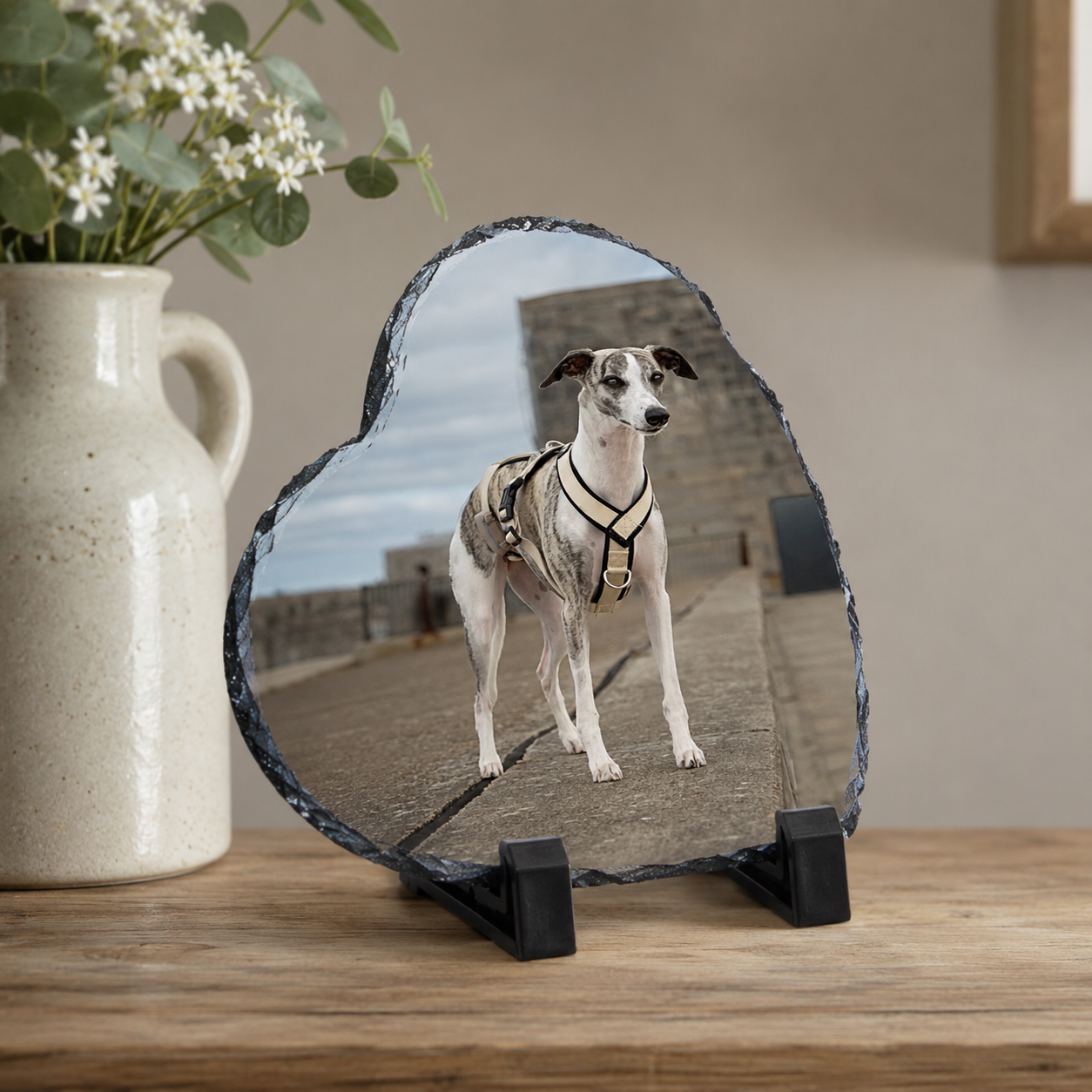 A heart-shaped slate photo plaque on a black stand, featuring a printed image of an Italian Greyhound dog wearing a harness, displayed on a wooden table next to a ceramic vase.