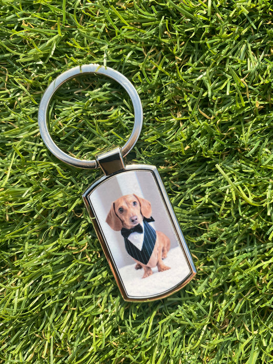 A custom rectangular metal keychain with a silver-toned split ring, featuring a printed photo of a dachshund wearing a suit, placed on a green grass background.