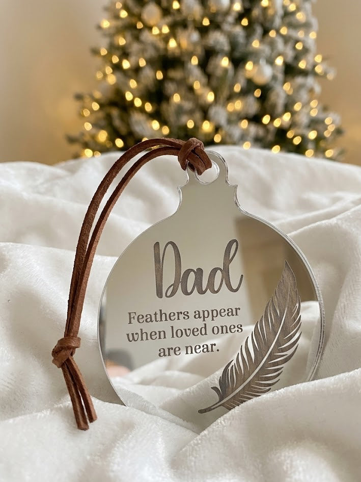 A round silver-toned ornament with a brown leather hanging loop, engraved with the word 'Dad' and a feather, resting on white fabric with a blurred Christmas tree in the background.