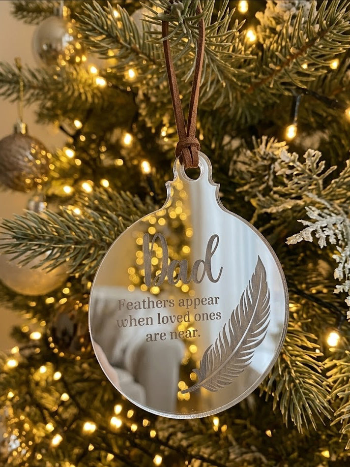 A round clear acrylic Christmas ornament hanging from a brown ribbon on a decorated Christmas tree. The ornament features the word 'Dad' in a large script font, with the phrase 'Feathers appear when loved ones are near.