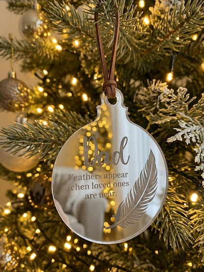 A round clear acrylic Christmas ornament hanging from a brown ribbon on a decorated Christmas tree. The ornament features the word 'Dad' in a large script font, with the phrase 'Feathers appear when loved ones are near.